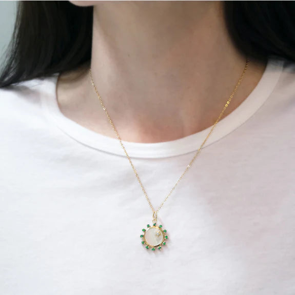 Front of mini Happiness sun charm on 18K gold chain, mother-of-pearl center with emerald and diamond accents, worn on model
