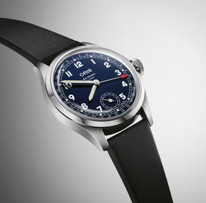 Oris Big Crown watch with a silver stainless steel case, a dark blue dial with white Arabic numerals and hands, and a black textured leather strap, shown on a light gray gradient background.