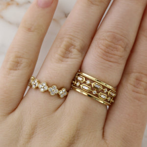 Two gold rings on a hand: a thin floral diamond band on the ring finger and a wide geometric diamond band on the middle finger