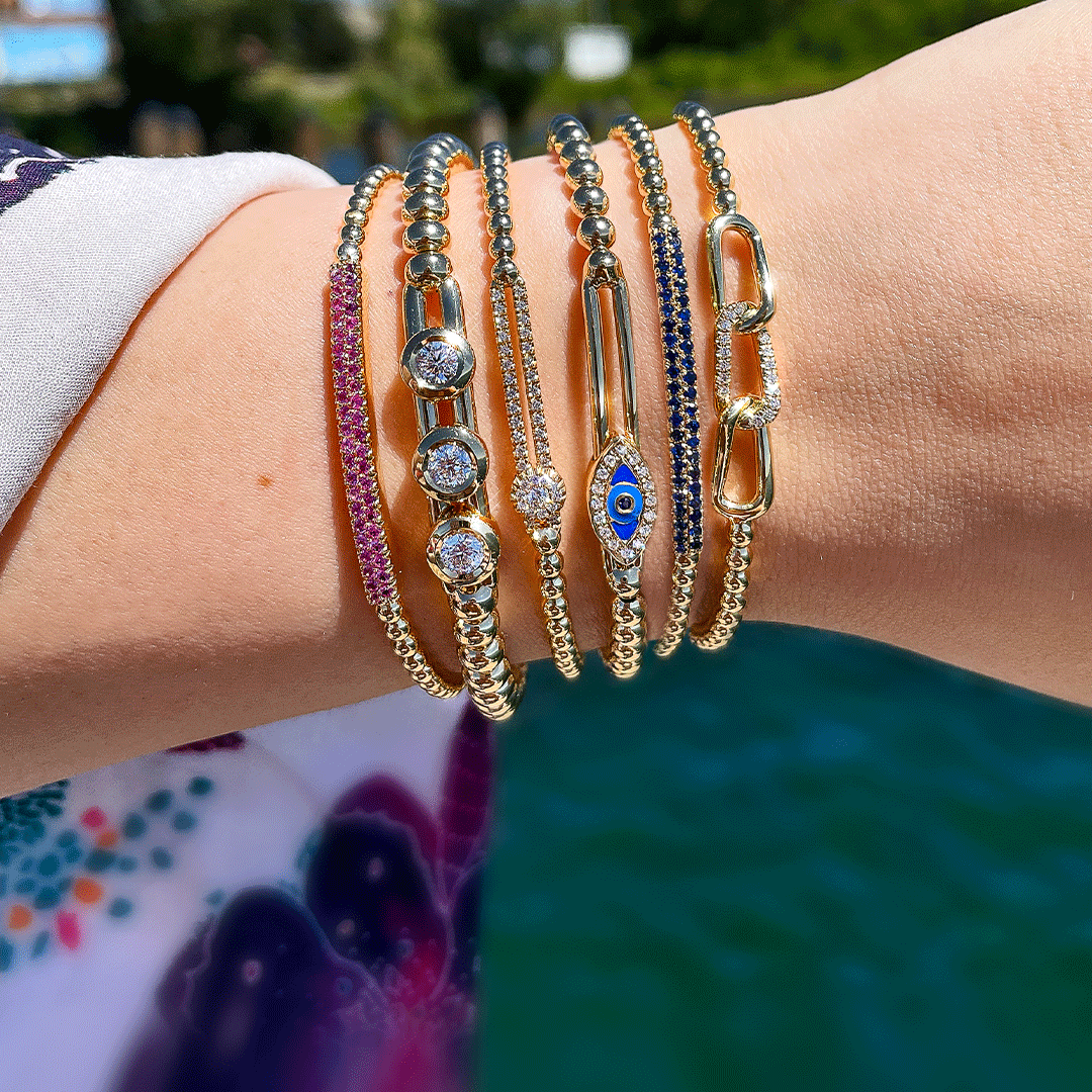 Seven gold beaded bracelets: pink pavé band, three-stone diamonds, blue evil-eye charm, chain-link accent; wrist over water