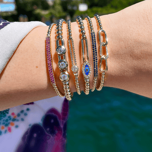 Seven gold beaded bracelets: pink pavé band, three-stone diamonds, blue evil-eye charm, chain-link accent; wrist over water