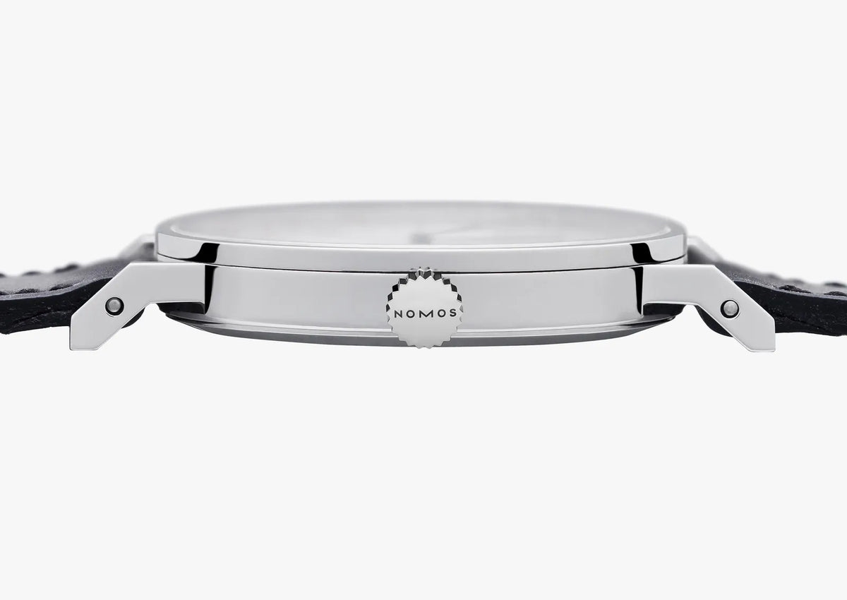 Close-up side view of a silver-toned wristwatch with a black strap, showing the side lugs and a small circular dial detail marked NOMOS on a light gray background.