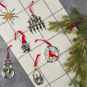 Metal holiday ornaments on checked cloth: gnome with lights, cardinal disk, star, snowman, evergreen sprig, red ribbons.