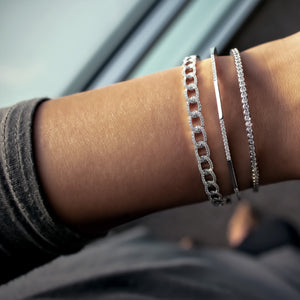 Three silver bracelets (chain, slim bar, and tennis styles) on a wrist beside a window.