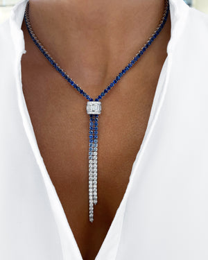 A close-up of a luxury Y-necklace worn by a model. The necklace features a central square-cut diamond pendant with three cascading strands: one in deep blue sapphires, one in clear cubic zirconia, and one in silver-toned metal.