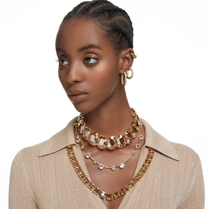 Model wearing gold-tone Dextera medium hoop earrings with white pavé crystals, styled with layered necklaces on white background