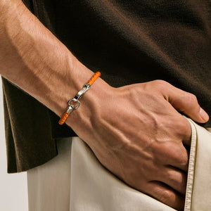 A close-up of a man's wrist wearing a bright orange beaded bracelet with a silver-toned clasp, set against a dark brown shirt and light beige trousers.