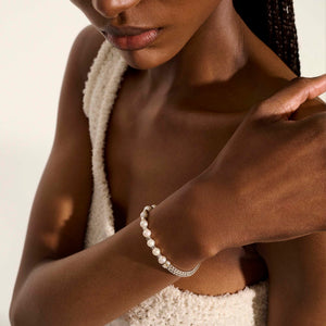 a lady wearing a white top with her hand on her shoulder with a pearl and silver bracelet on.