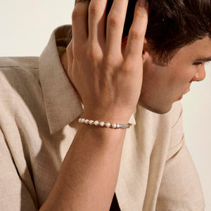 a man wearing a pearl and silver bracelet with his hand on his head wearing a beige shirt.