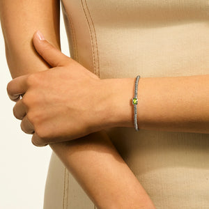 A close-up of a person's wrist wearing a delicate silver chain bracelet with a small light green square gemstone at the center, set against a beige dress.