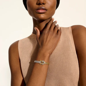 A close-up of a model wearing a delicate double-band bracelet with a central diamond-encrusted oval clasp, one band in yellow gold and the other in white gold or platinum. The model is wearing a beige ribbed knit top against a solid off-white background.