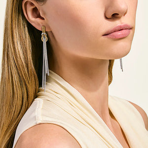 A close-up of a woman wearing a long dangling earring with a gold-toned knot top and multiple silver-toned chain drops, paired with a cream draped top against a light neutral background.
