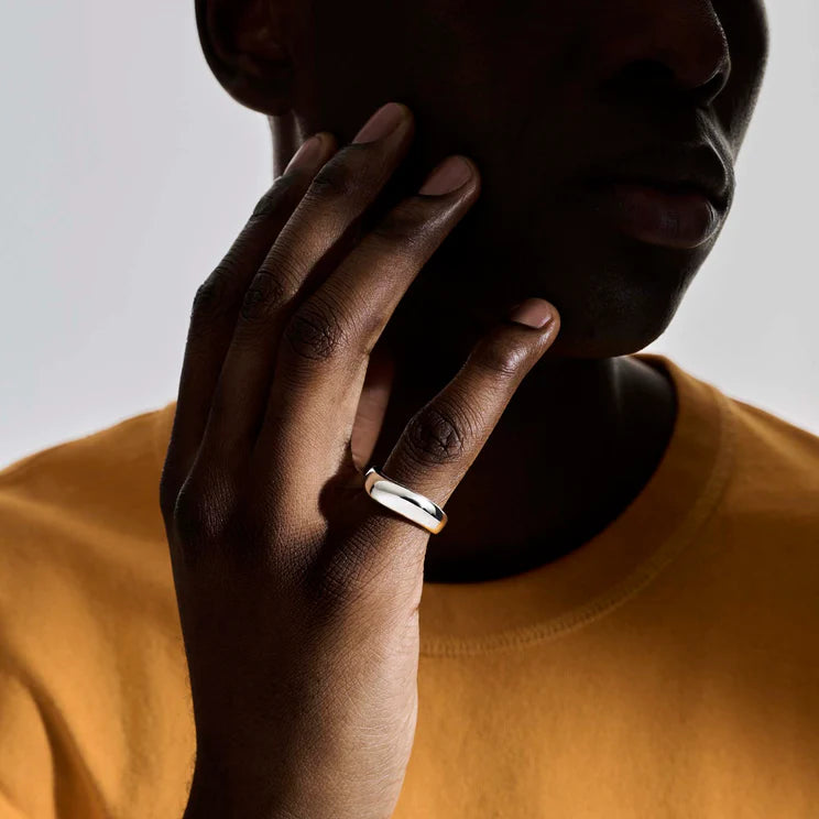 A close-up lifestyle shot of a model wearing a minimalist silver band ring on their ring finger, dressed in a mustard yellow t-shirt against a neutral gray background.