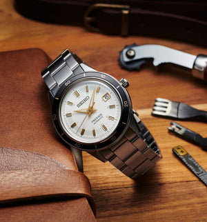 Three-quarter view of Seiko SRPG03 Presage '60s steel watch with light beige dial, black bezel, date window on leather background