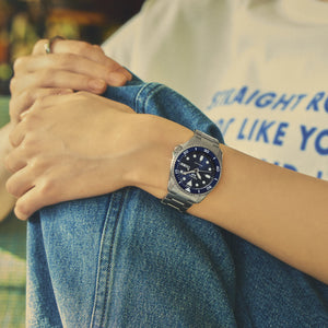 A silver stainless steel wristwatch with a dark blue dial and white markers, worn on a person's wrist. The person is wearing a white graphic t-shirt and blue denim jeans, with their hands crossed over their lap.