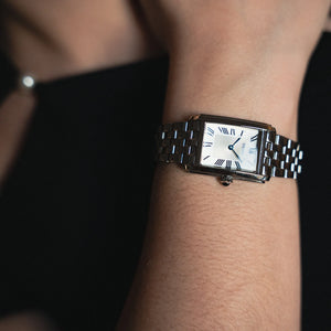 A close-up of a wrist wearing a rectangular stainless steel watch with a white dial, blue Roman numeral hour markers, and blue hands, set against a dark background.