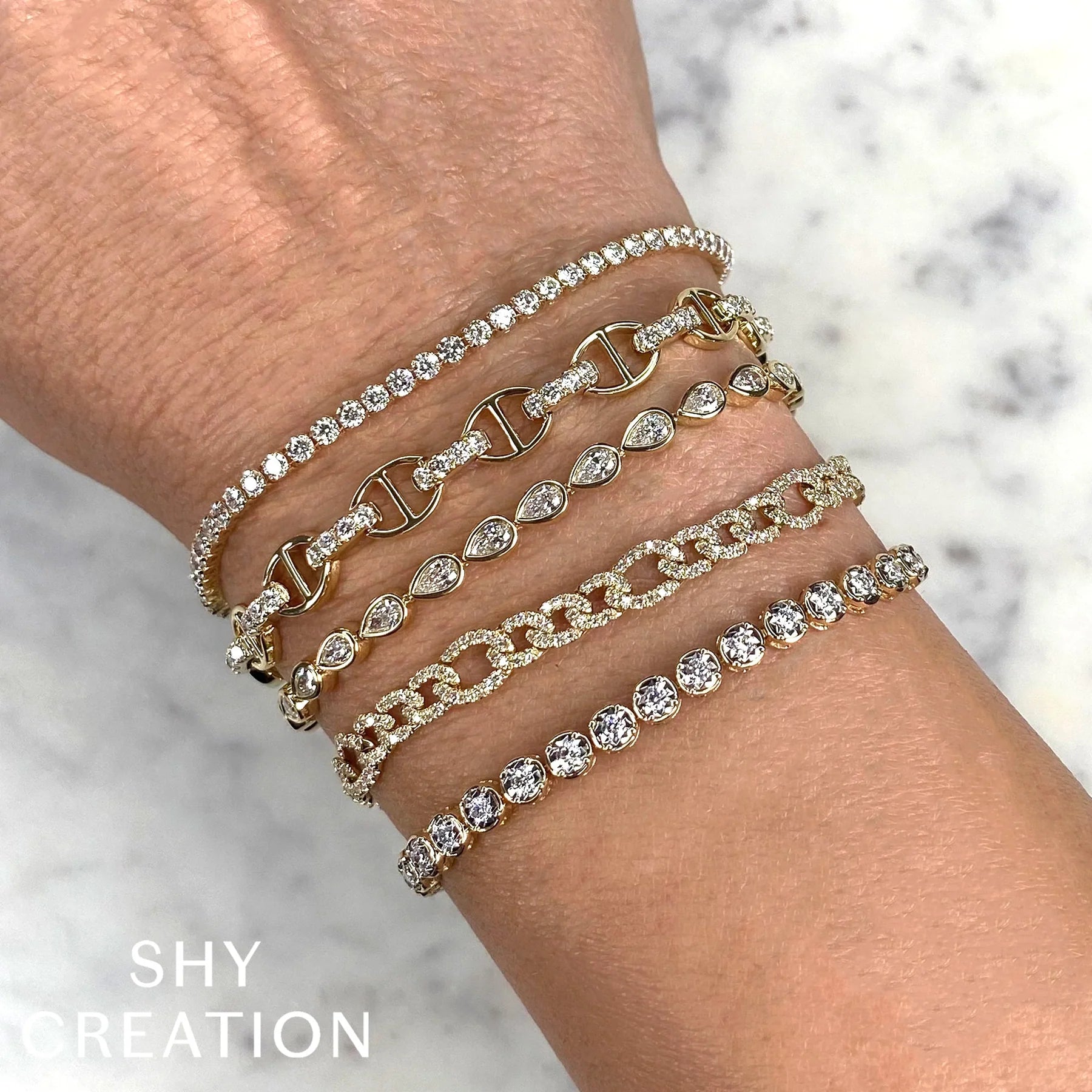 Stack of six gold diamond bracelets on a wrist with mixed link and teardrop settings; SHY CREATION visible