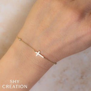 Wrist view of 14K yellow gold cross bracelet with 0.03 ctw diamond bezel and delicate chain on light marble background