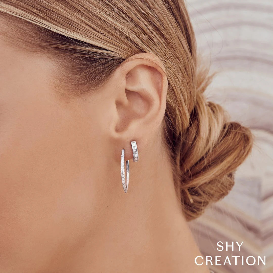 Close-up of a woman's left ear wearing two silver diamond hoop earrings; brand logo SHY CREATION visible.