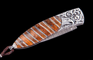 A close-up of a decorative knife or tool handle with a carved silver-toned metal cap featuring an eagle motif, a striped brown and silver inlay body, and a dark cord loop, shown against a solid black background.