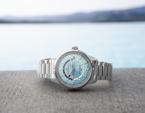 Front view of Freedom 60 GMT 40mm watch with blue dial, red-tip GMT hand, date at 3 o'clock, stainless bracelet and blurred sea
