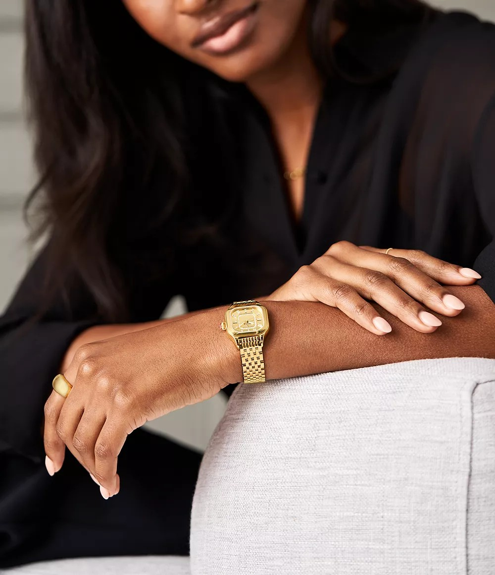 Close-up of Meggie gold-plated watch on a model’s wrist showing diamond-accented dial and gold bracelet on a light gray chair