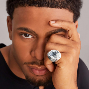 Close-up of hand wearing sterling silver signet ring with raised skull motif and textured sides against neutral background