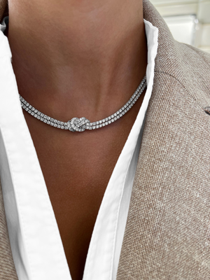 A close-up of a model wearing a silver-toned diamond choker necklace with a knot design at the center, styled with a white collared shirt and a beige blazer.