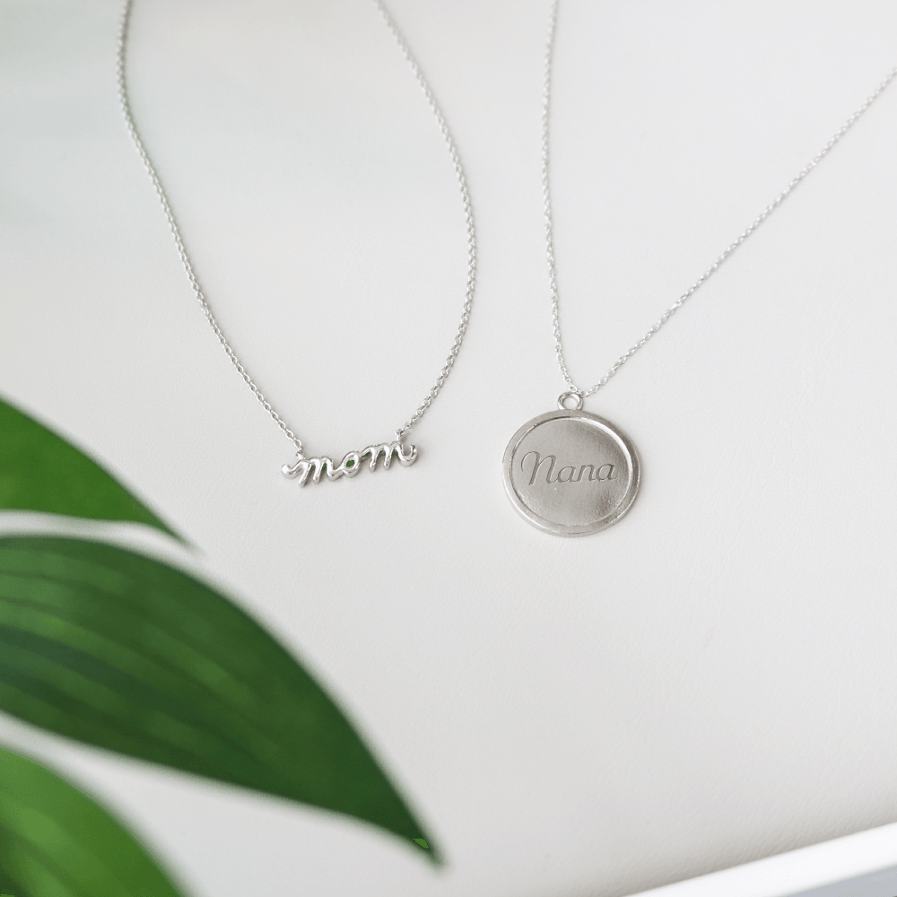 Two silver necklaces on white background: a script 'mom' pendant and a round engraved 'Nana' medallion, with a green leaf.
