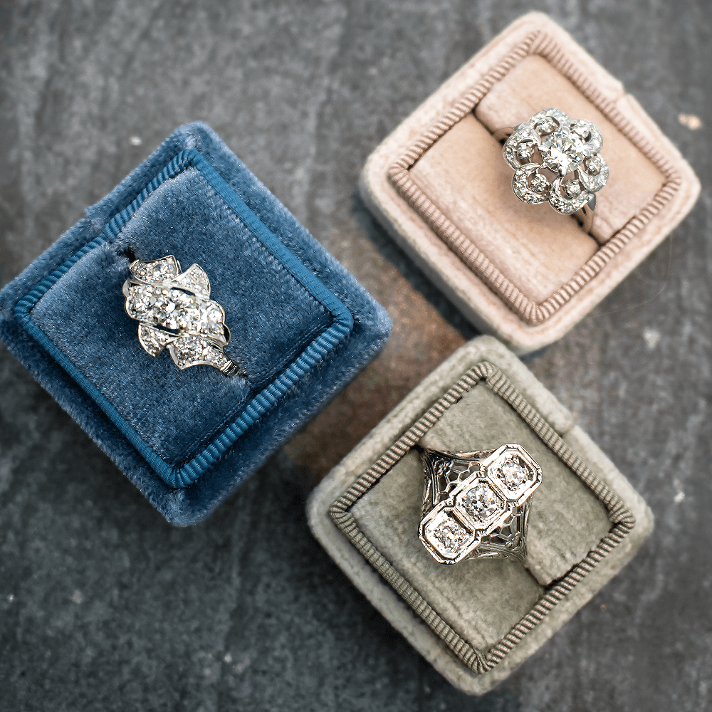 Three vintage diamond engagement rings displayed in blue, blush, and gray velvet ring boxes on dark slate.
