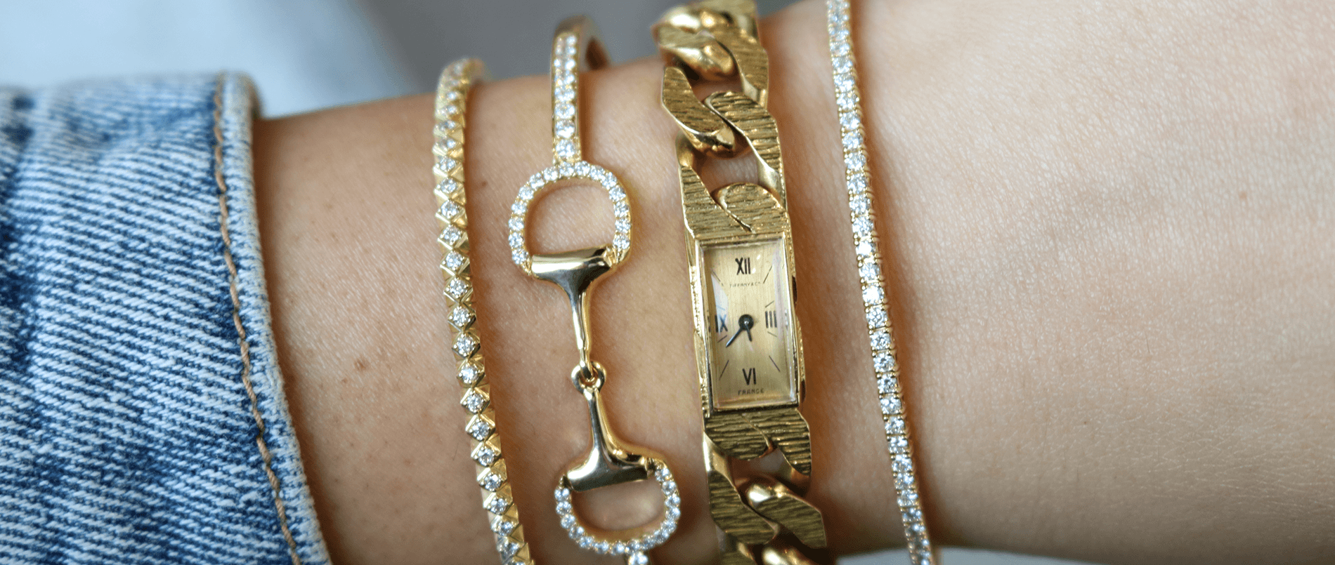 Wrist wearing stacked gold bracelets and a slim rectangular gold watch face showing XII III VI FRANCE, beside a denim cuff