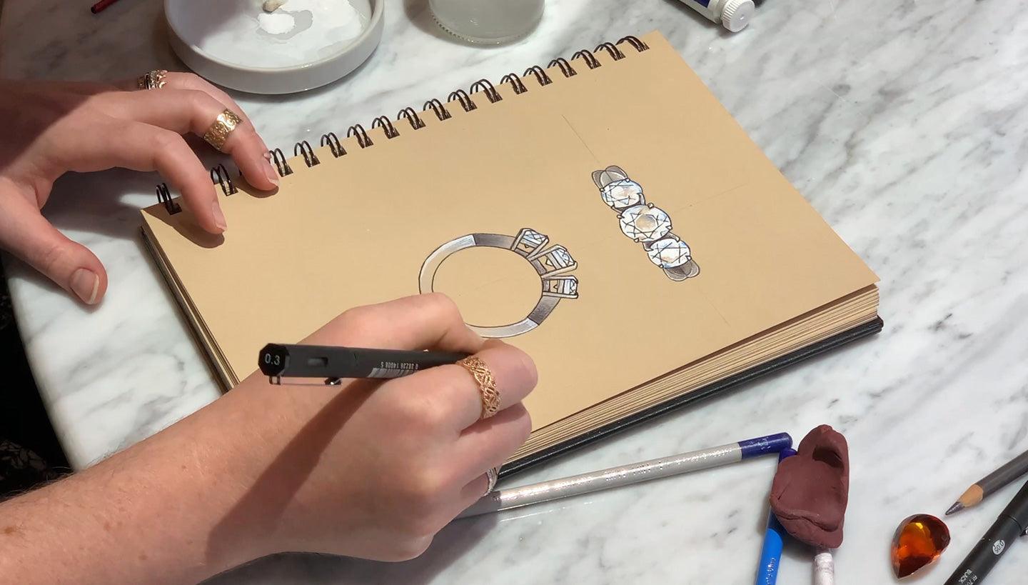 Hands sketching ring and diamond designs in a spiral sketchbook on a marble table; pen labeled "0.3"