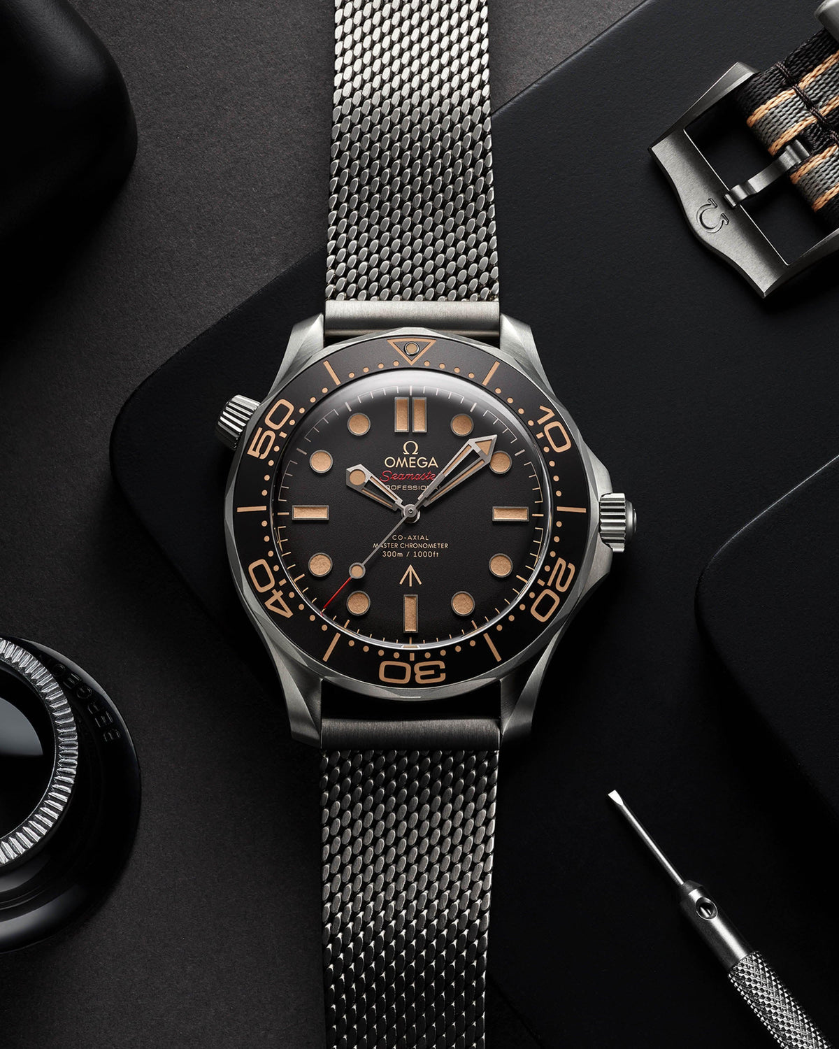 Close-up top-down of Omega Seamaster watch showing black dial with bronze markers and mesh bracelet on a dark matte background