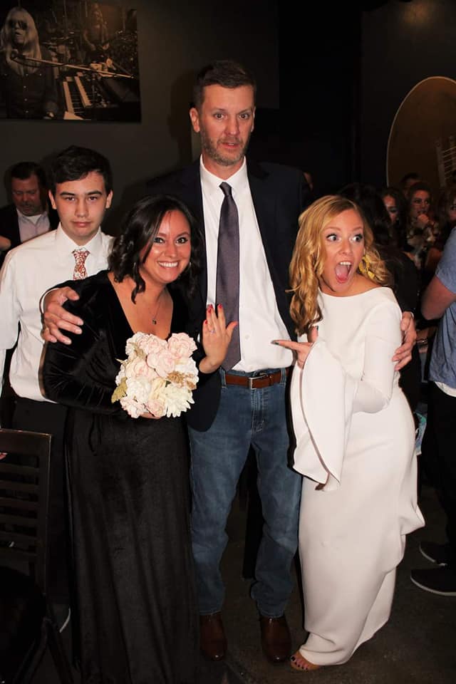 Bride in white gown pointing excitedly, groom in blazer and jeans, and bridesmaid in black holding a bouquet at a reception - Paiton & Jim
