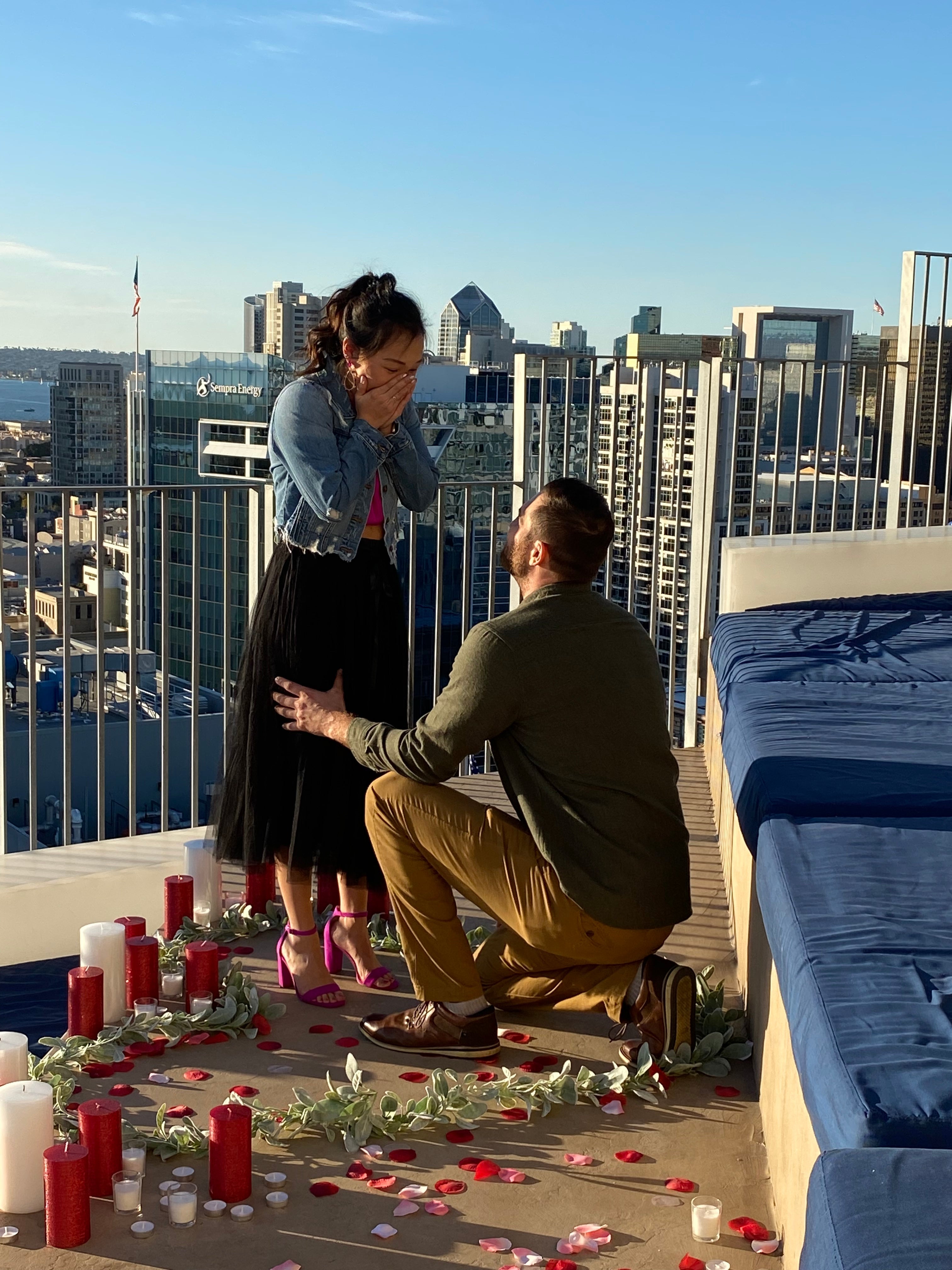 Ali and Joe: rooftop proposal with candles and rose petals; city skyline and 'Sempra Energy' building visible.