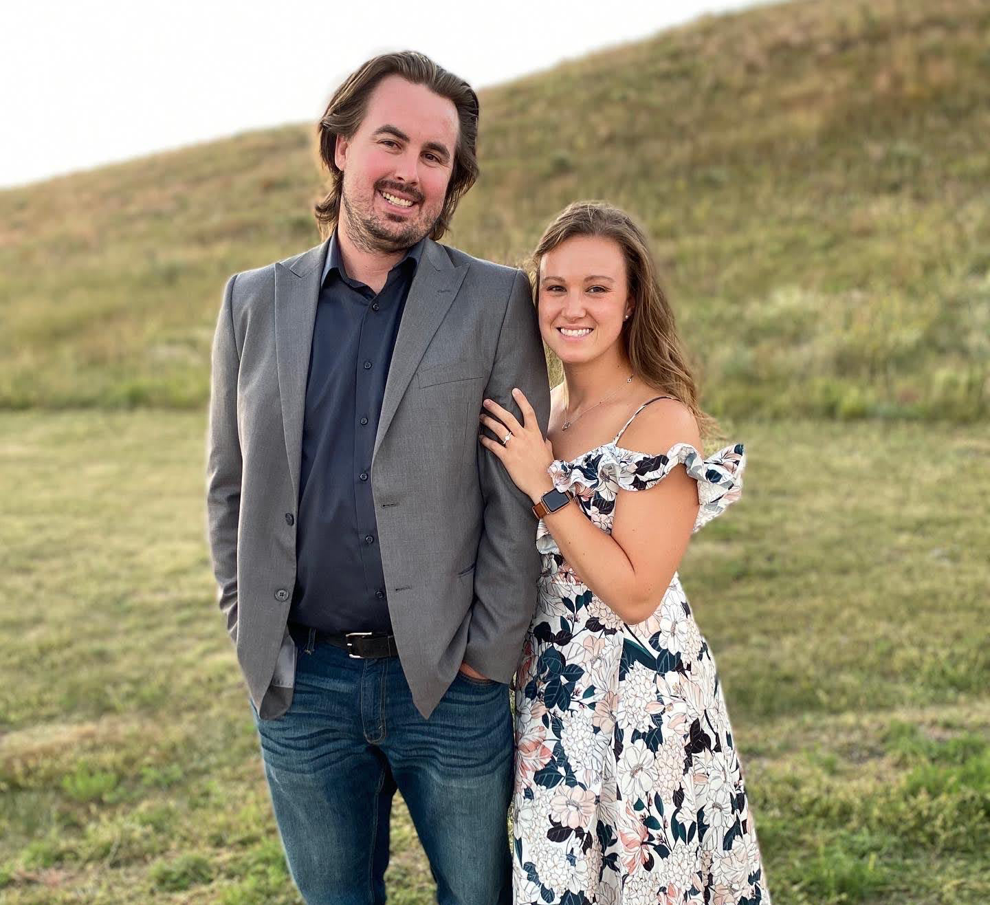 Michelle and Grant standing arm-in-arm in a grassy field; Michelle resting her left hand on Grant's arm showing a ring