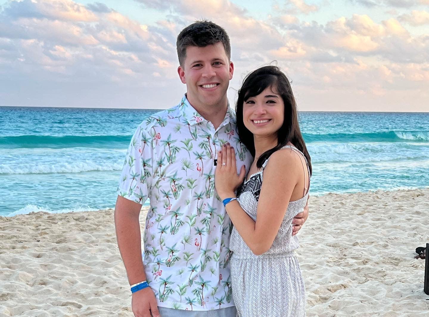 Smiling couple embracing on a sandy beach at sunset, woman showing engagement ring on left hand