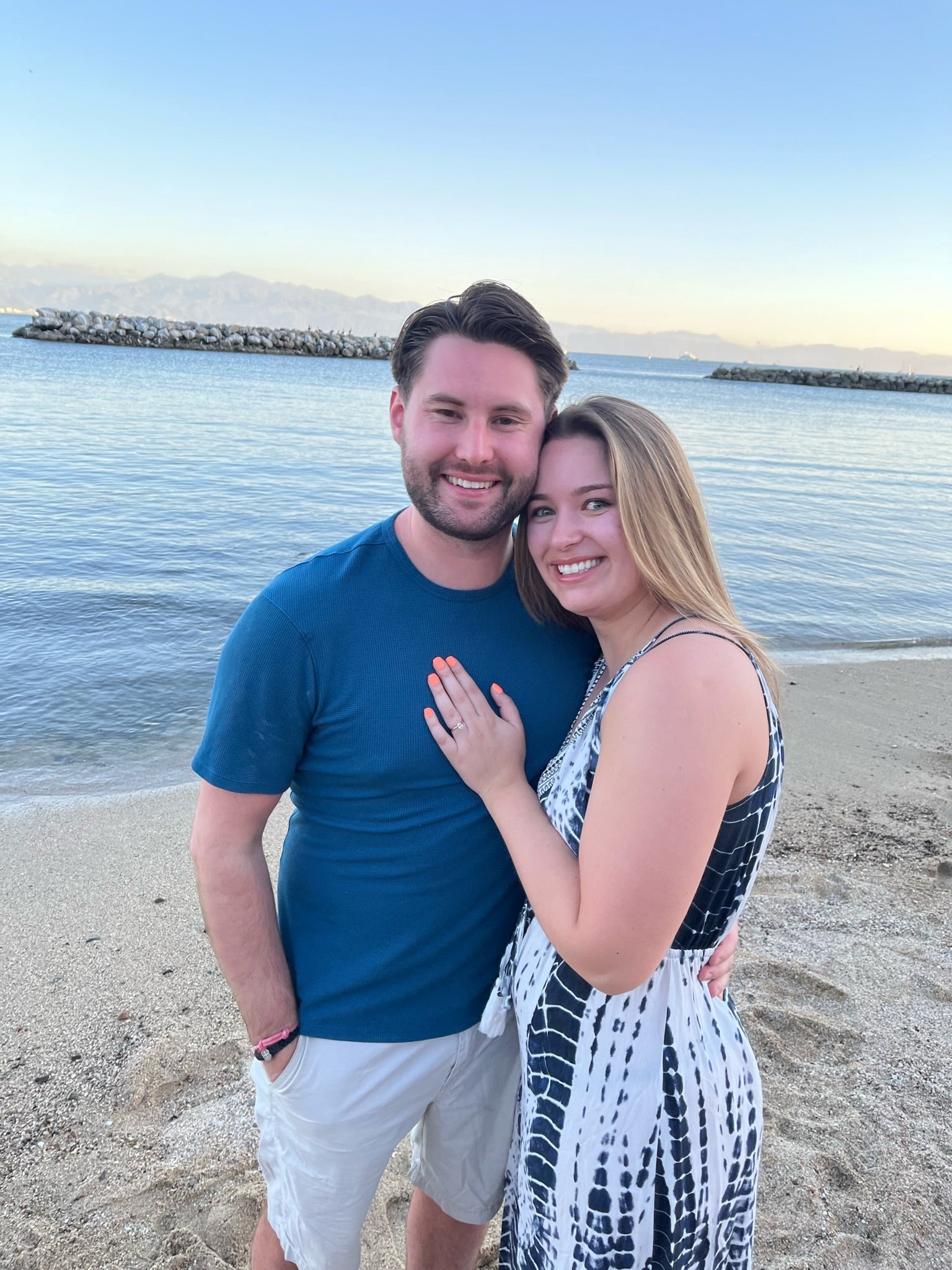 Maizie and Brady smiling on a beach at sunset; Maizie’s hand shows an engagement ring.