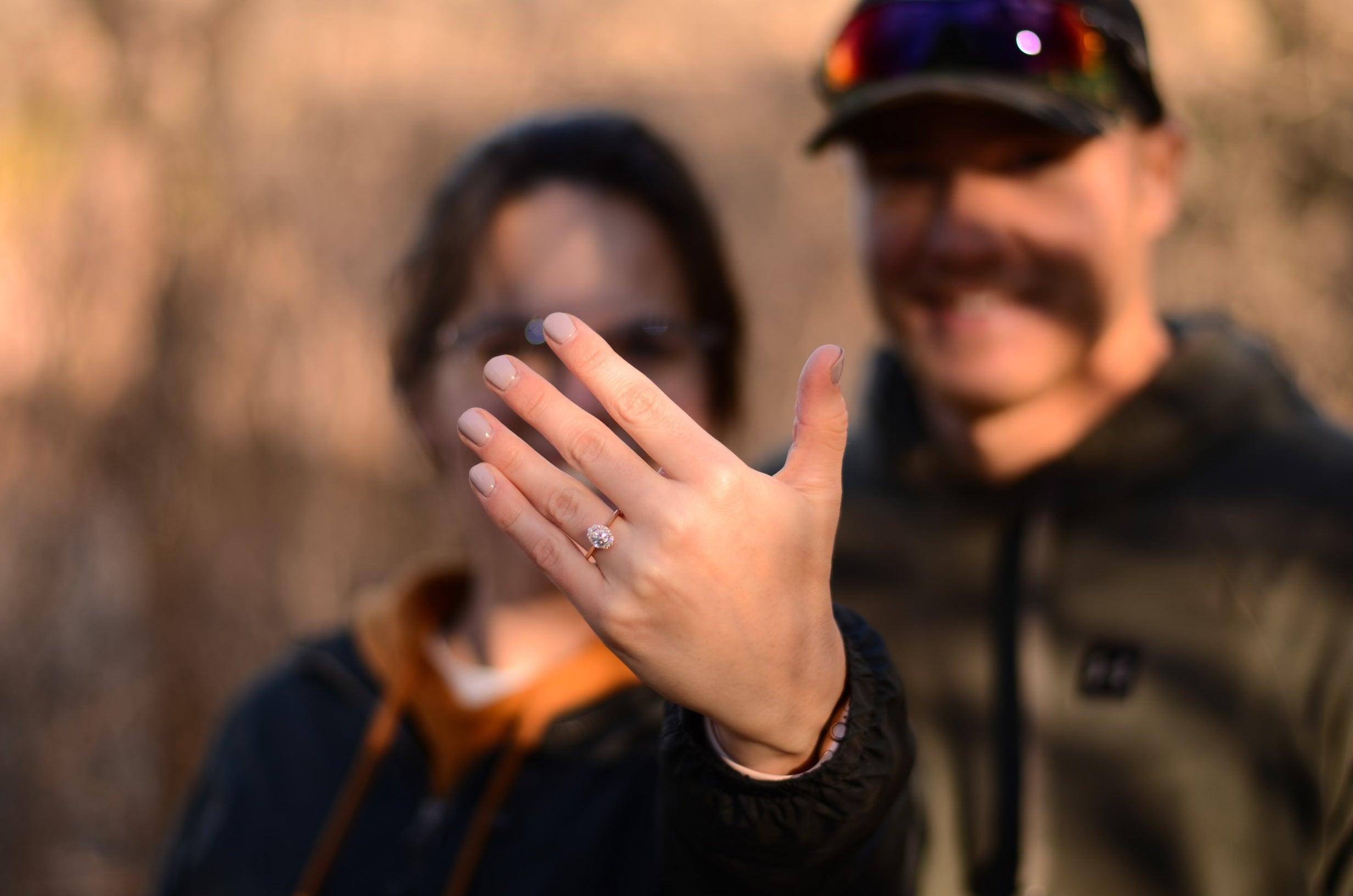 Woman showing engagement ring on left hand, partner smiling blurred behind — Jordan and Ian.