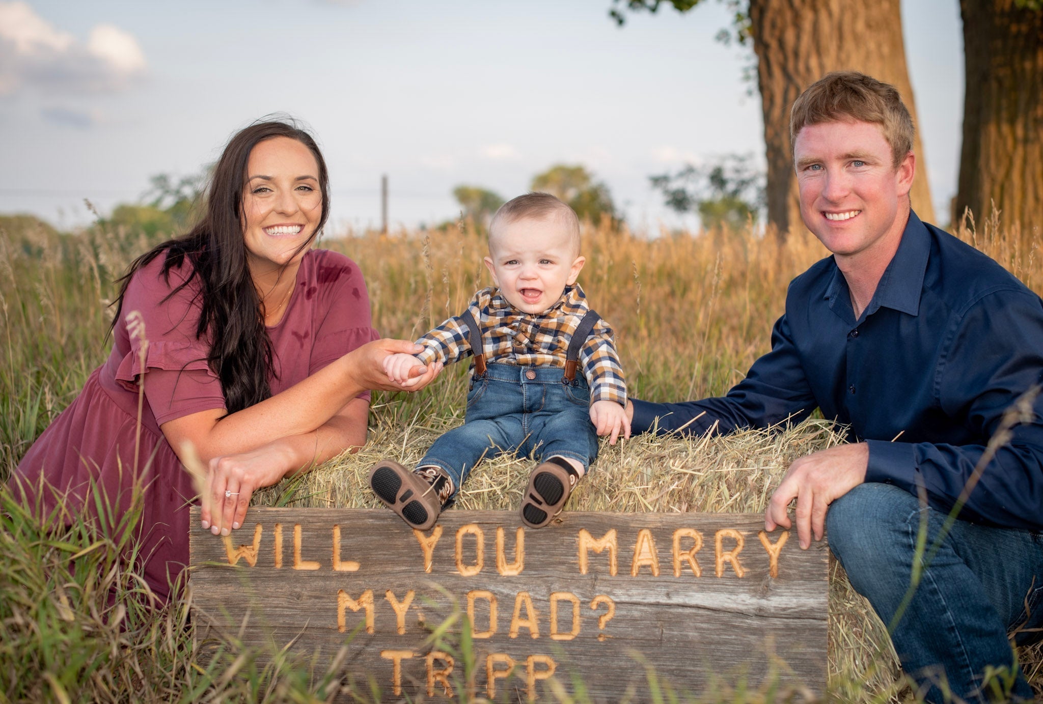 Devan and Troy with toddler on hay holding wooden sign reading WILL YOU MARRY MY DAD? TRIPP