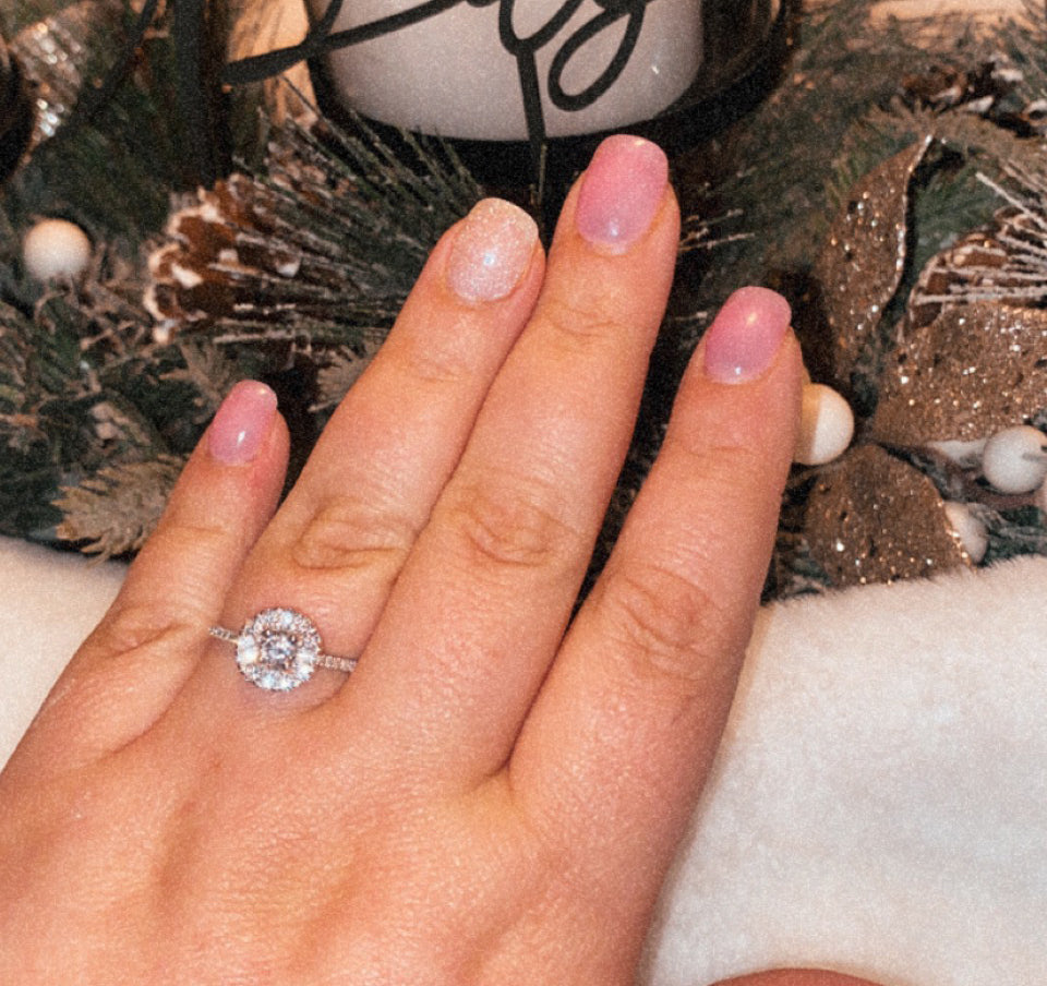 Left hand wearing halo diamond engagement ring with pink manicure over holiday decor; ornament reads Joy - Sidney & Brandon