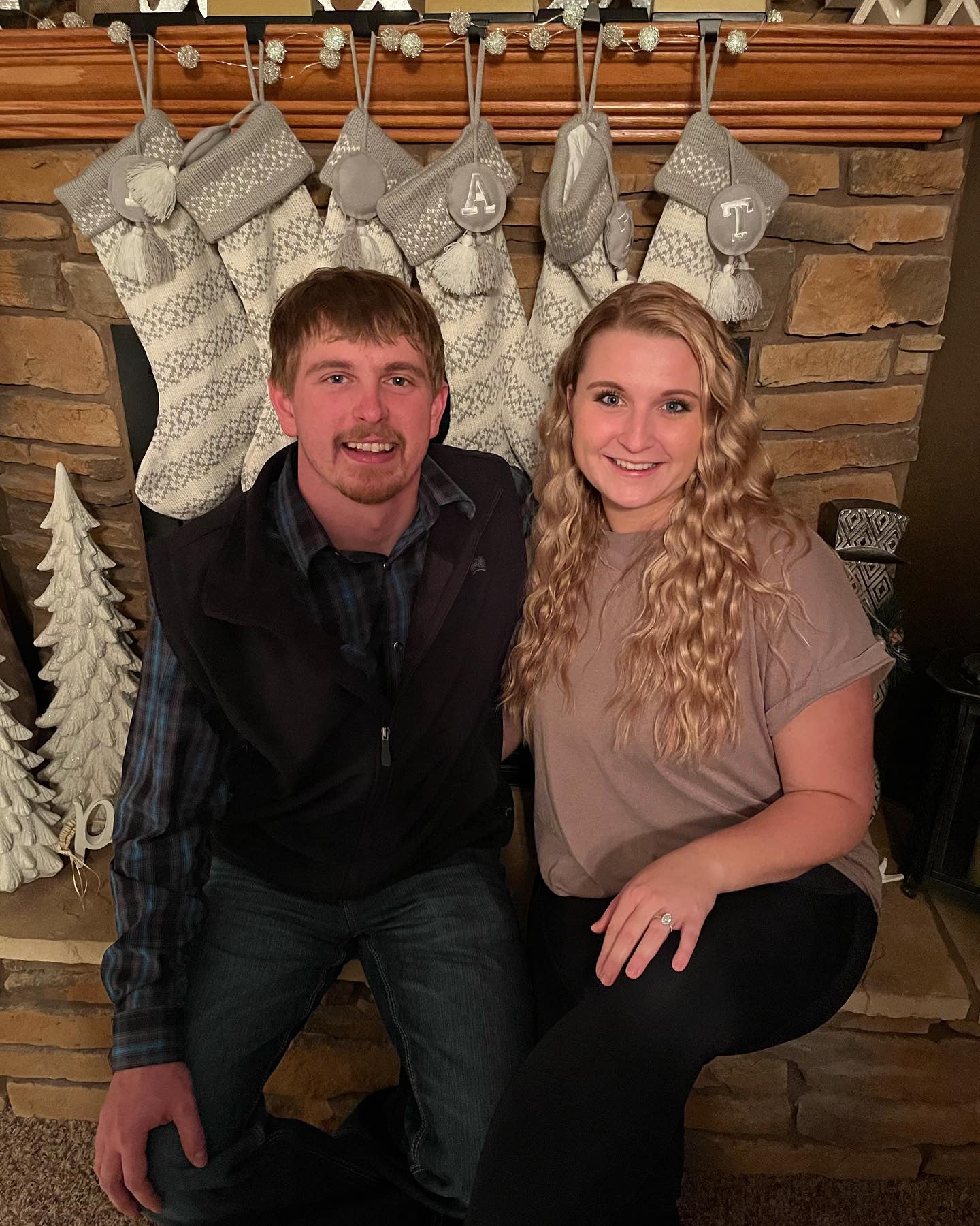 JaeCee and Zach seated by a fireplace with monogram stockings A and T; JaeCee shows her engagement ring.