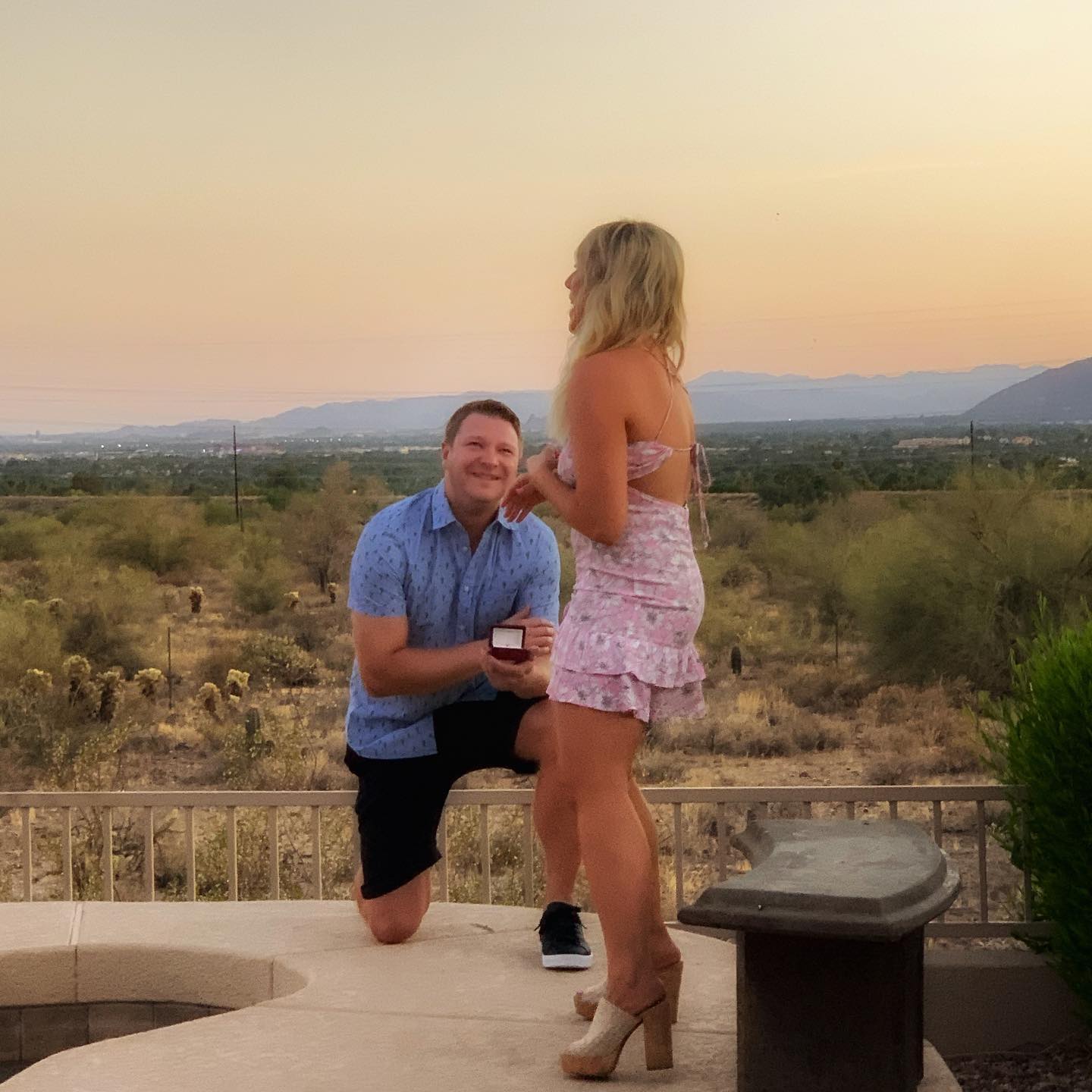 Matt kneeling and proposing with a ring box to Lexi on a patio at sunset, desert and mountains in the background.