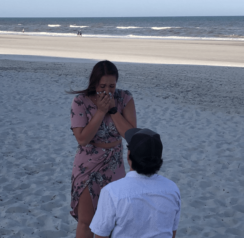 Hannah covering her mouth in surprise as Tommy kneels to propose on a sandy beach