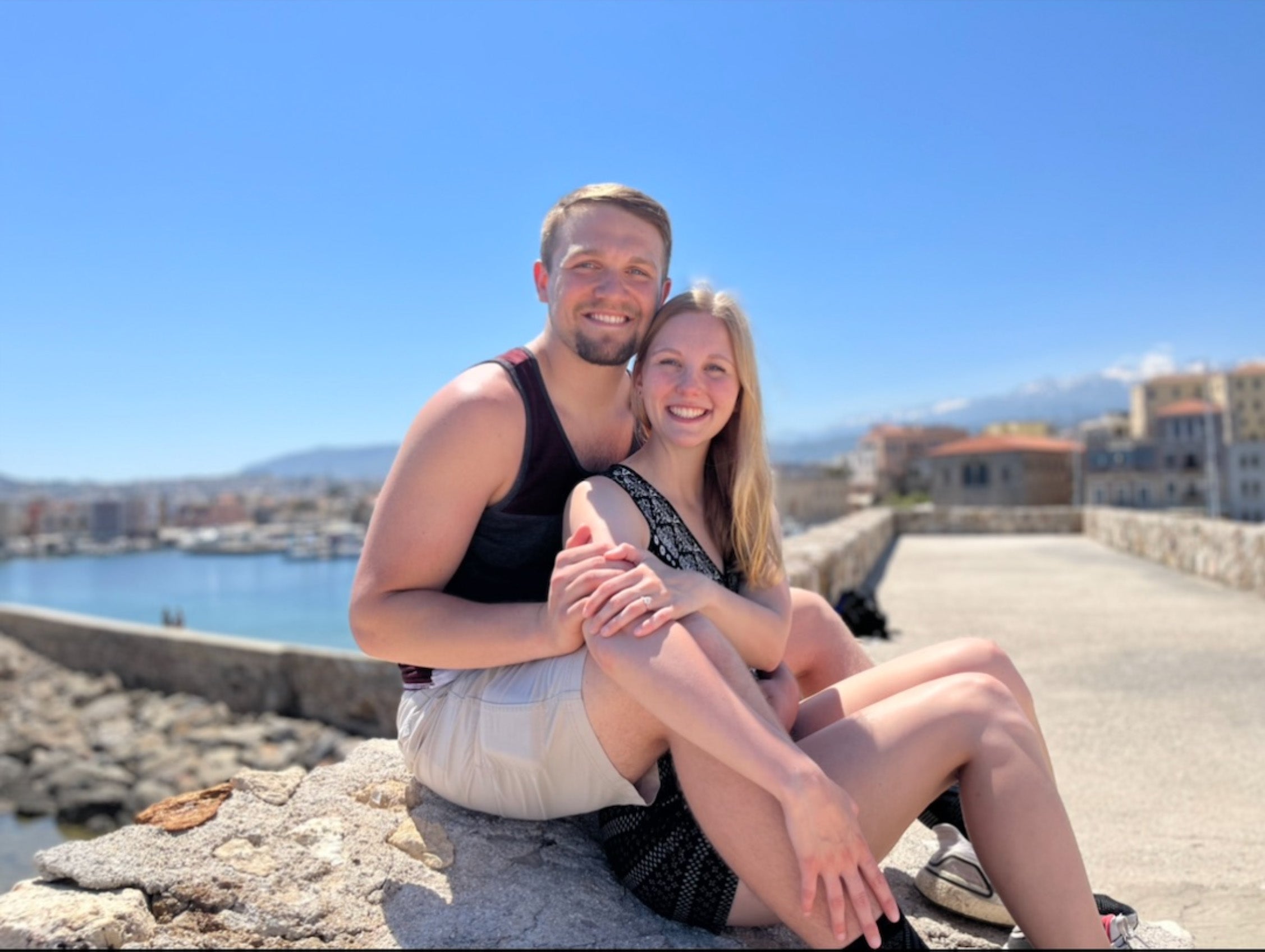 Brynn and Levi sitting on a stone seawall, smiling with harbor and buildings behind them