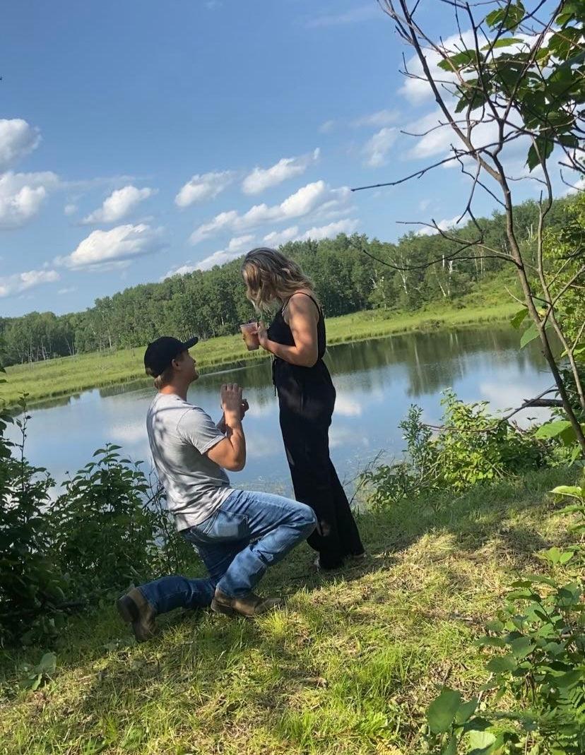 Ryan kneeling and proposing to Savanna beside a pond; she holds a drink and reacts.