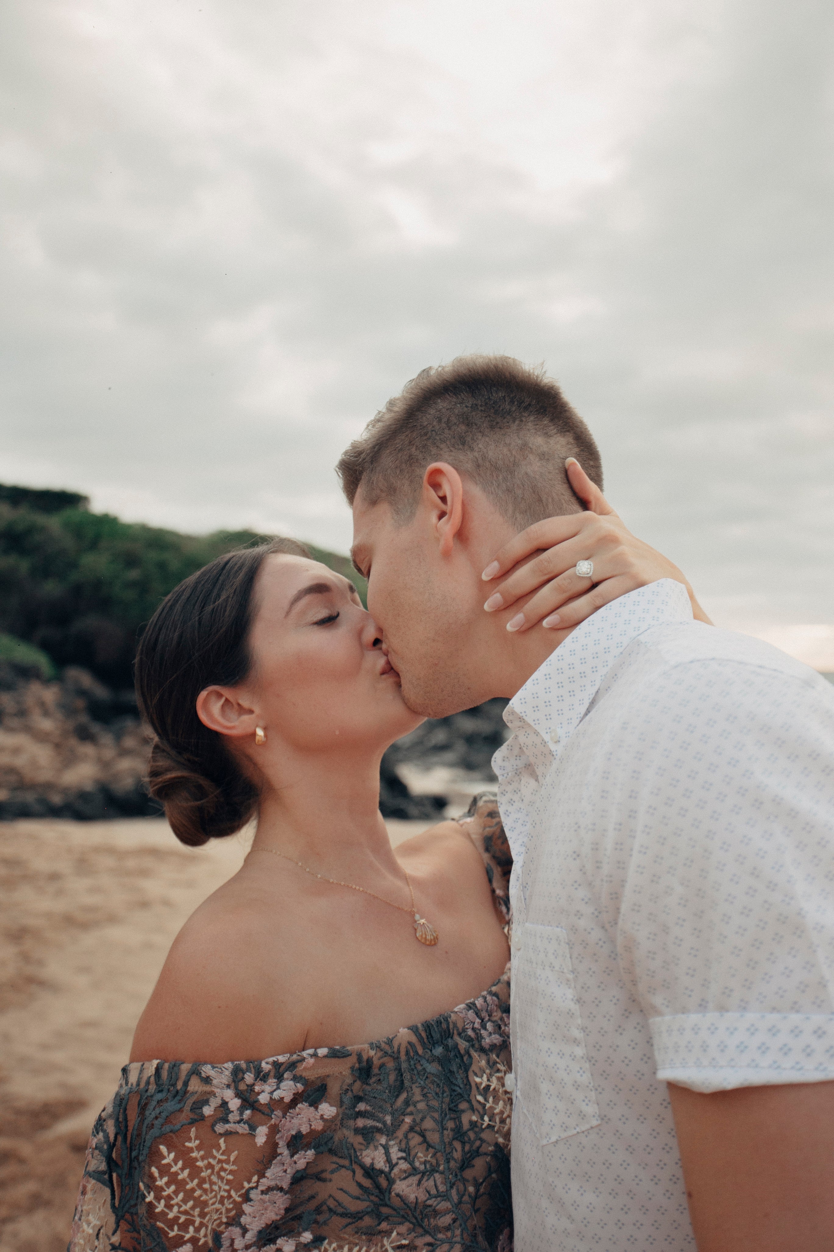 Lilliana and Luke kissing on a beach; Lilliana's hand with engagement ring rests on Luke's neck.