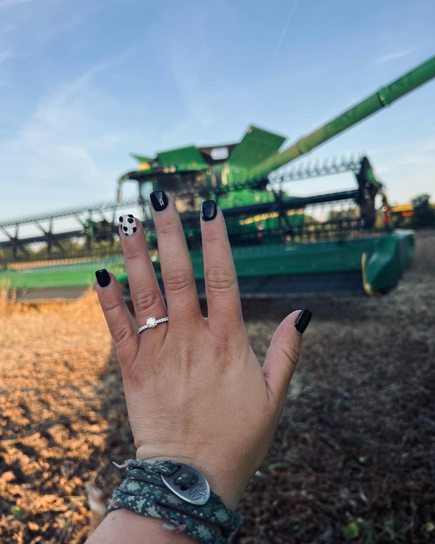 Hand with engagement ring and black cow-print manicure held up in front of a green combine harvester.