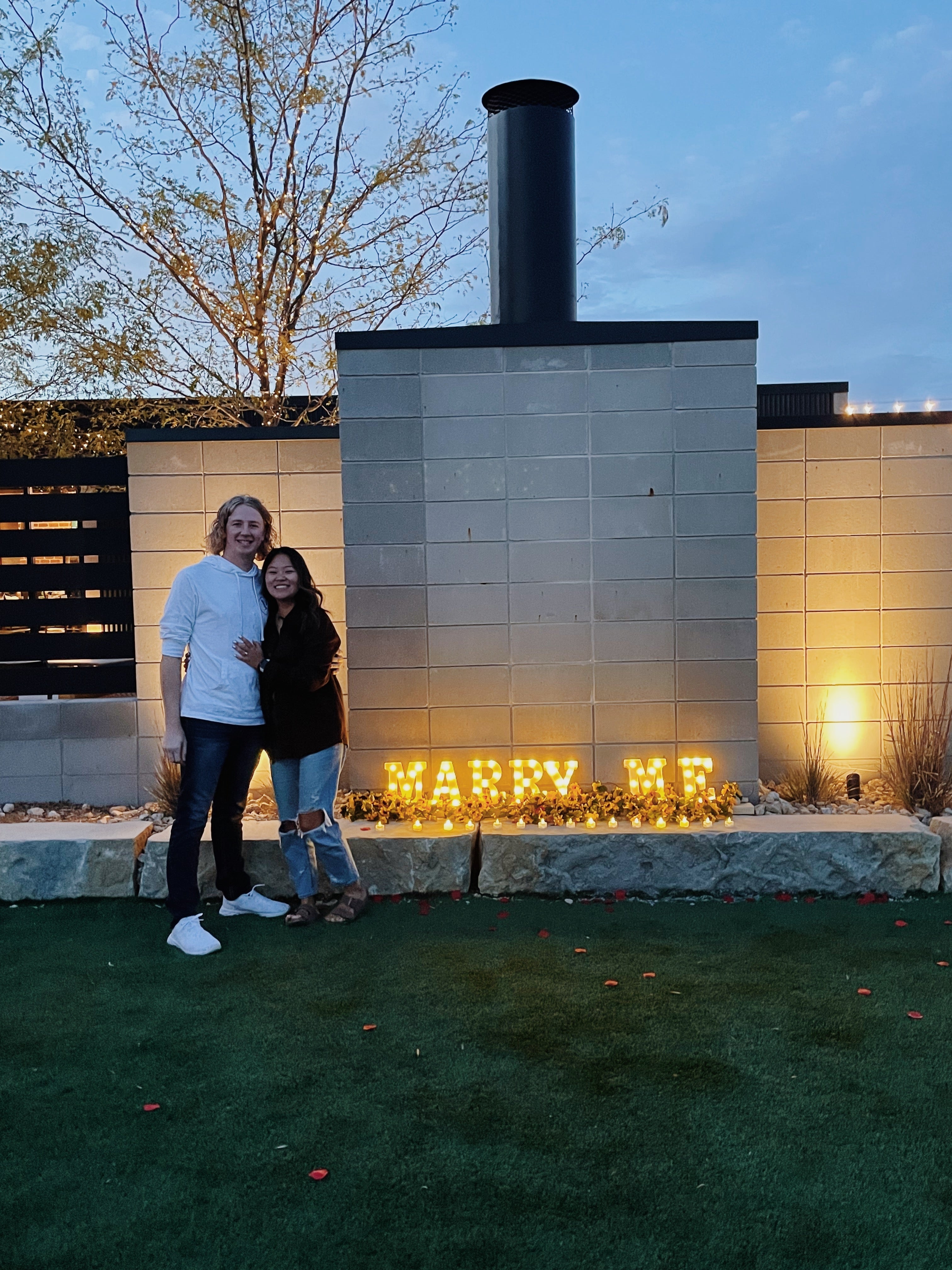 Amanda & Jeffrey embracing at dusk beside illuminated letters reading MARRY ME against a stone wall