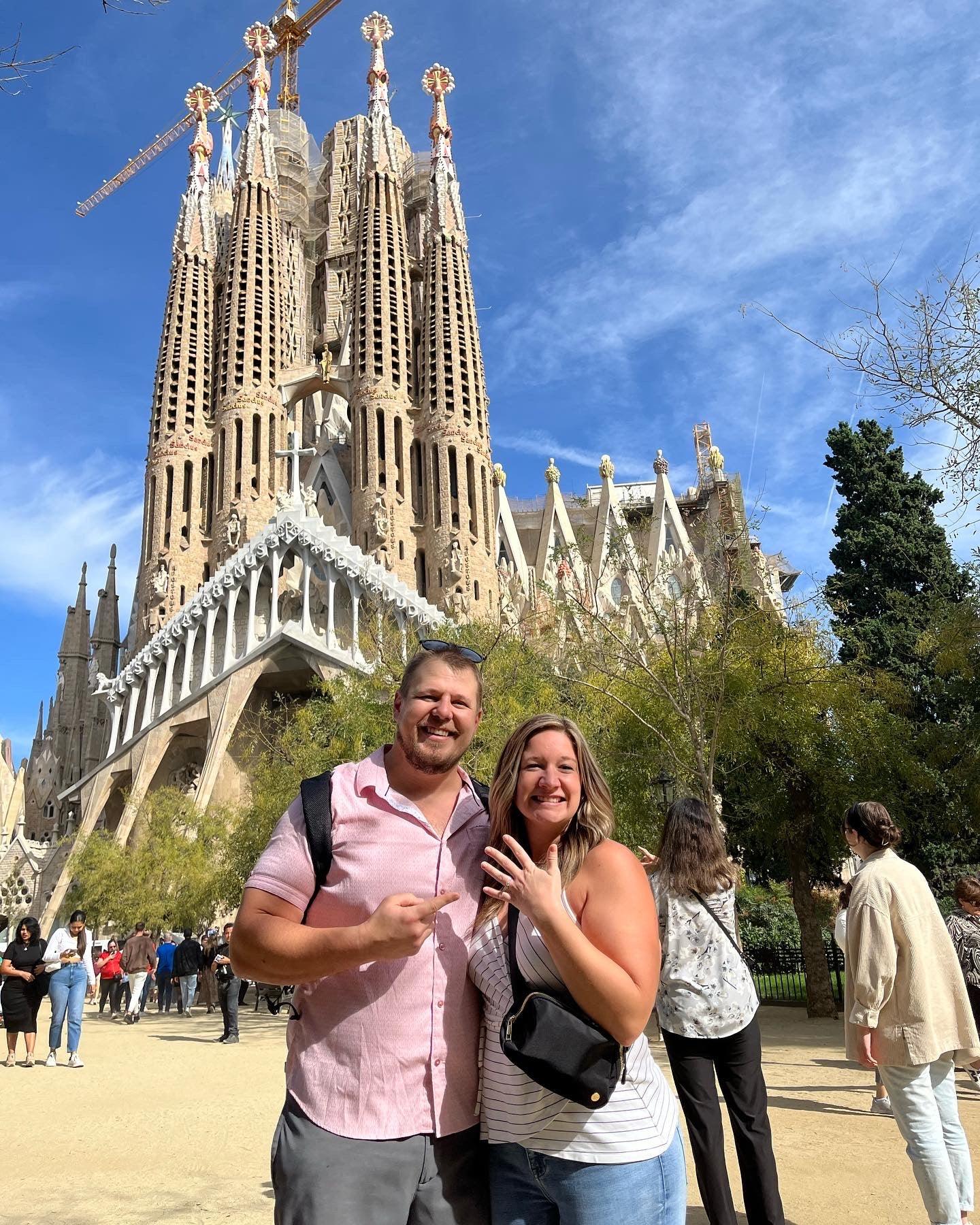 Smiling couple - Jenna & Tyler - showing engagement ring in front of La Sagrada Família, Barcelona